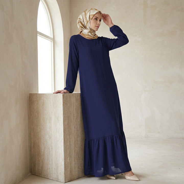 Woman in a long blue dress and headscarf standing in a softly lit room.