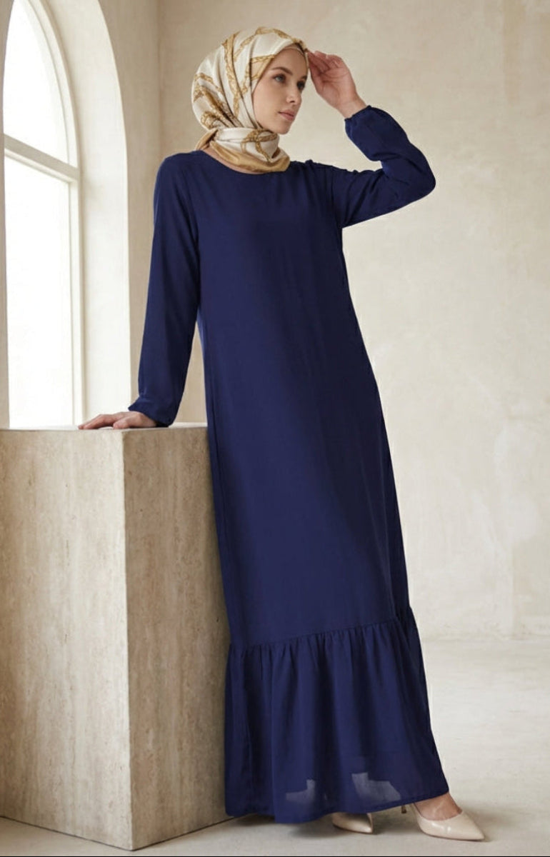 Woman in a long blue dress and headscarf standing in a softly lit room.