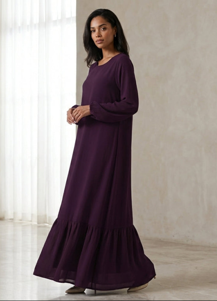 Woman in a long purple dress standing in a softly lit room with neutral walls and flooring.
