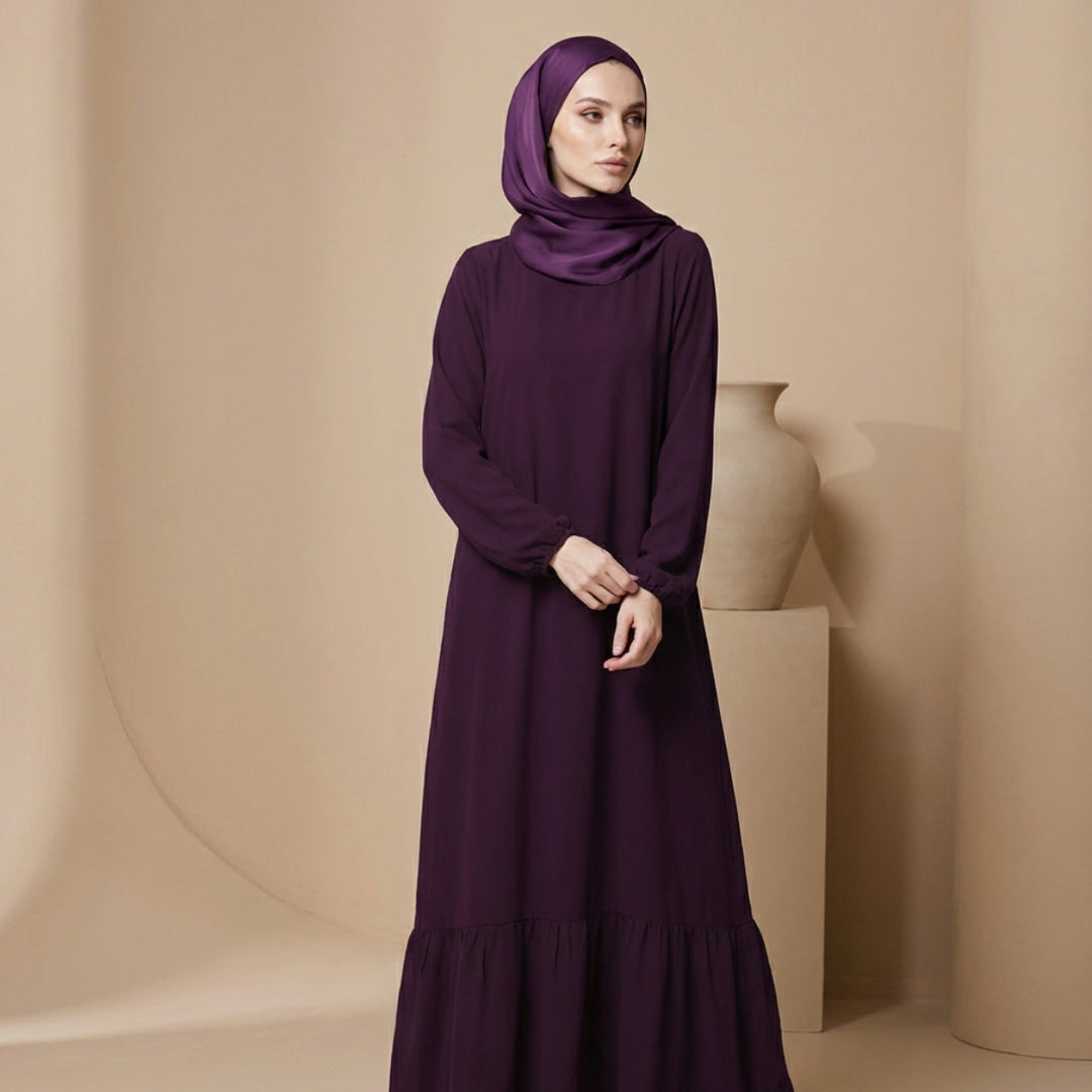 Woman wearing a long purple dress and hijab against a beige background