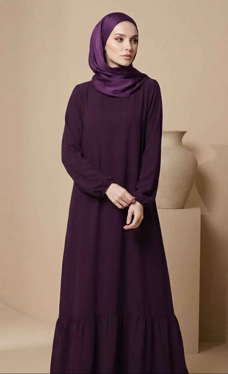 Woman wearing a long purple dress and hijab against a beige background