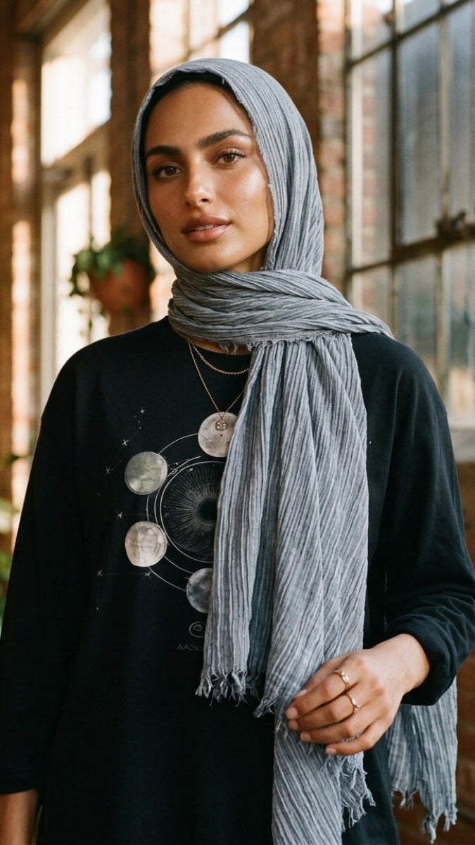 A woman wearing a light gray striped hijab and a black long-sleeve top with a moon phase graphic, standing in a brick-walled indoor space with warm sunlight.