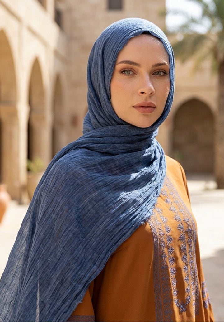 Woman wearing a blue hijab and brown top in an outdoor setting with arches and palm trees.