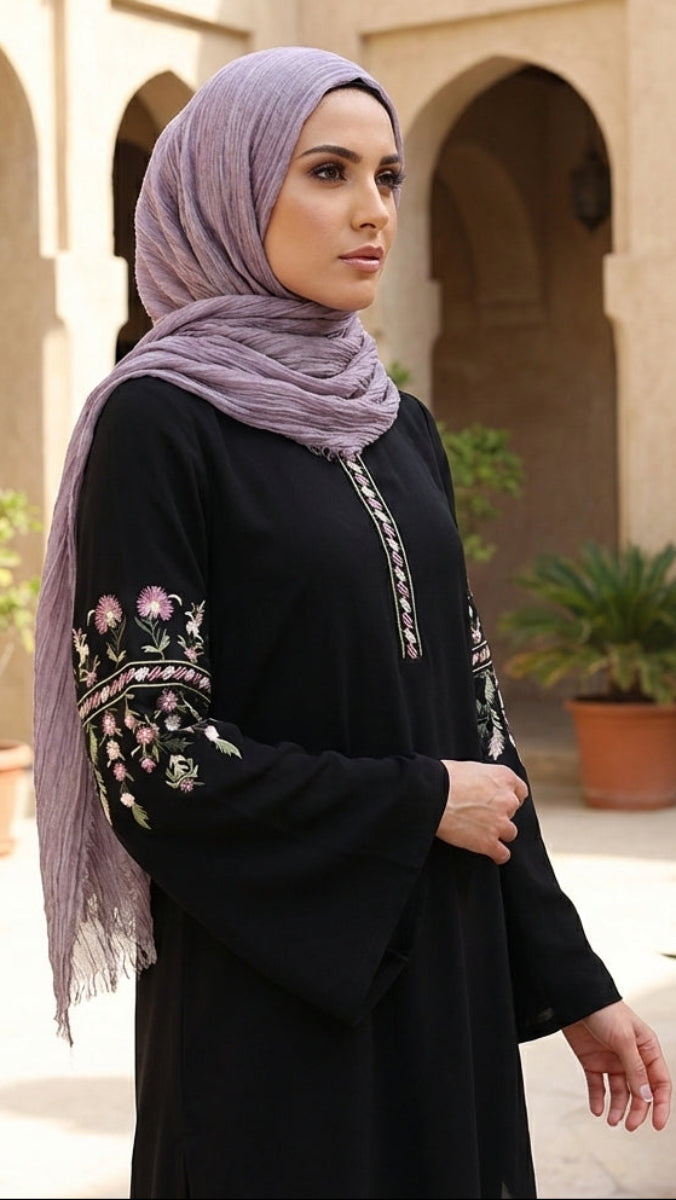 Woman wearing a black dress with embroidery and a light purple hijab in an outdoor setting with arches and plants.