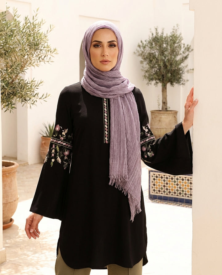 Woman wearing a black outfit with floral embroidery and a light purple hijab in an outdoor setting.