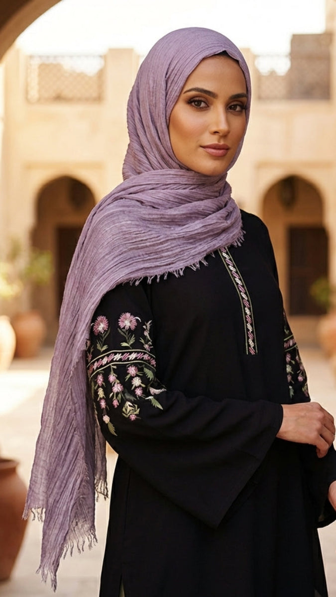 Woman wearing a black outfit with floral embroidery and a purple hijab in an outdoor setting.