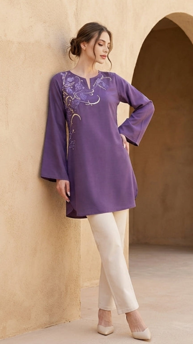 Woman in a purple embroidered top and white pants standing in front of a beige wall with an archway.