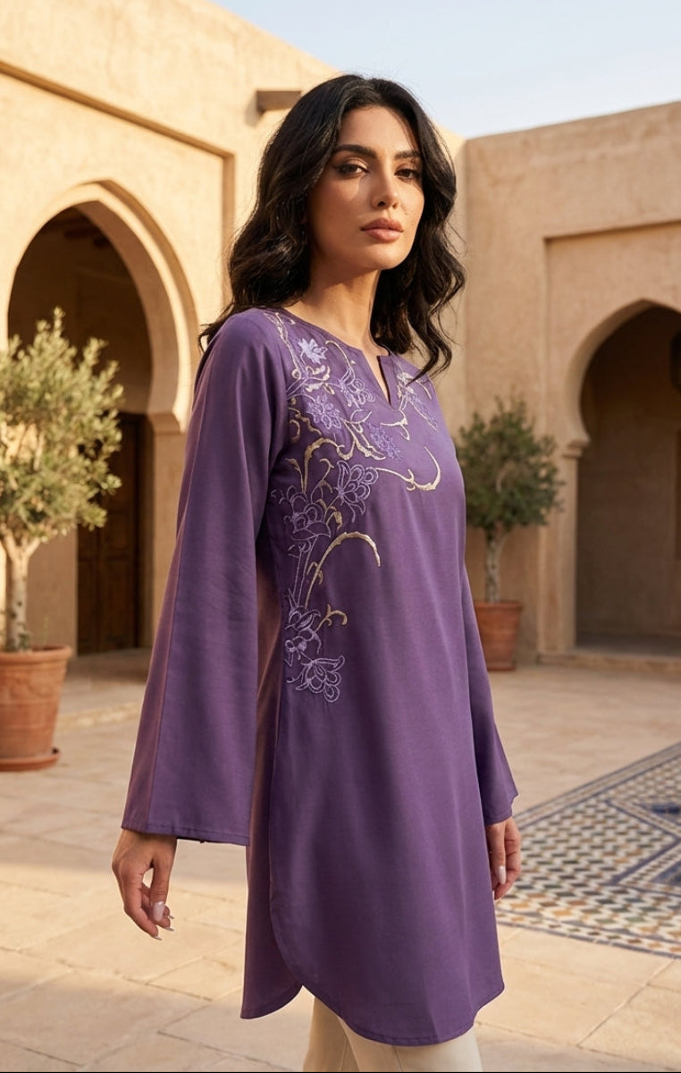 Woman wearing a purple embroidered dress standing in a courtyard with architectural elements and plants.