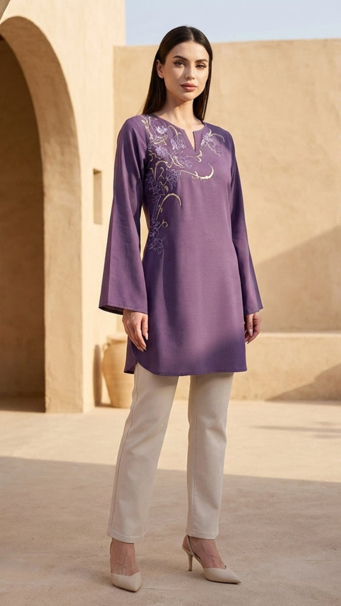 Woman wearing a purple embroidered top and white pants in an outdoor setting with beige walls and pots.