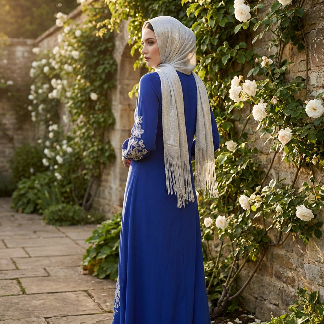 Woman in a blue dress with floral patterns standing in a garden with stone walls and white flowers.