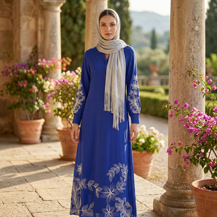 Woman in a blue dress with floral embroidery standing in a garden setting.