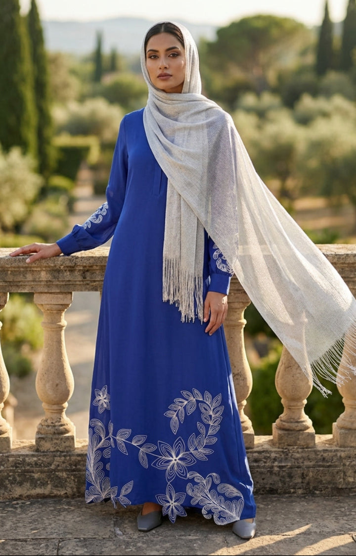 Woman in a blue dress with white embroidery and a white scarf standing on a stone balcony with a scenic background.
