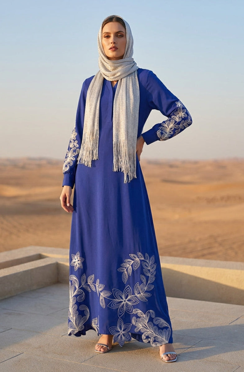 Woman in a blue dress with white embroidery and hijab standing in a desert landscape.