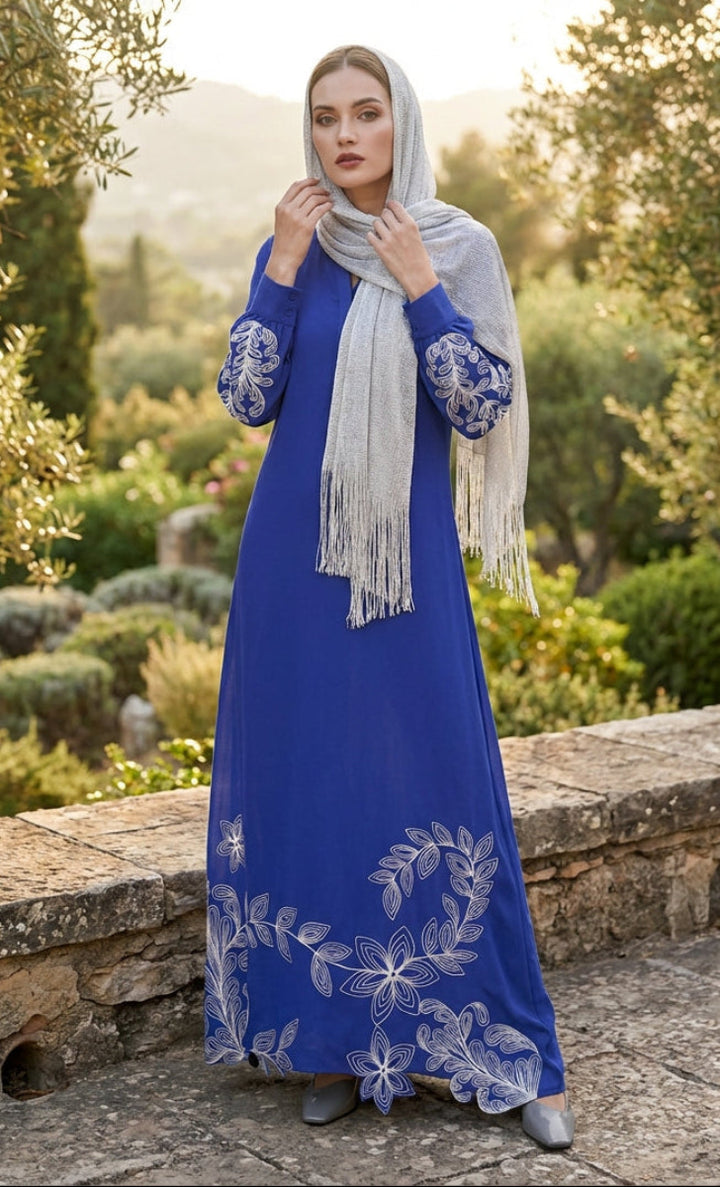 Woman in a blue dress with white embroidery standing in a garden.