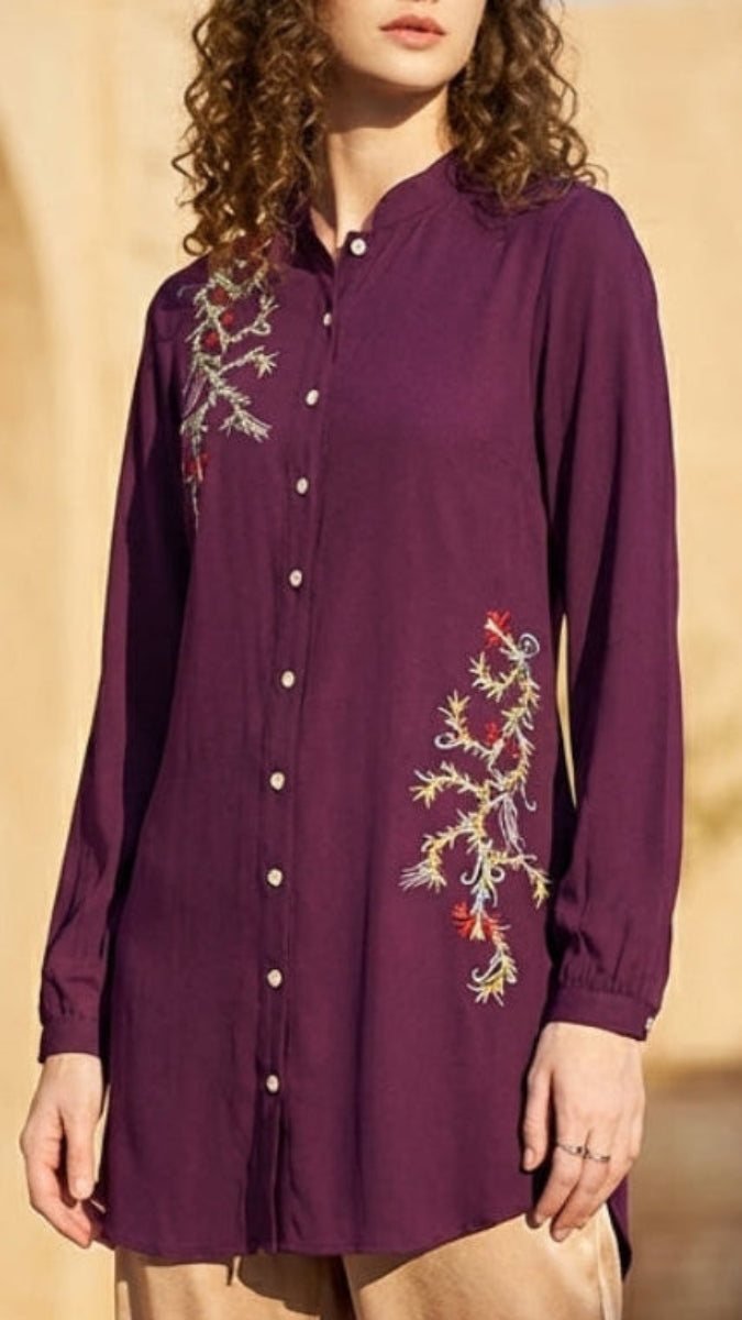 Woman wearing a purple embroidered shirt standing in front of a stone building with arches.