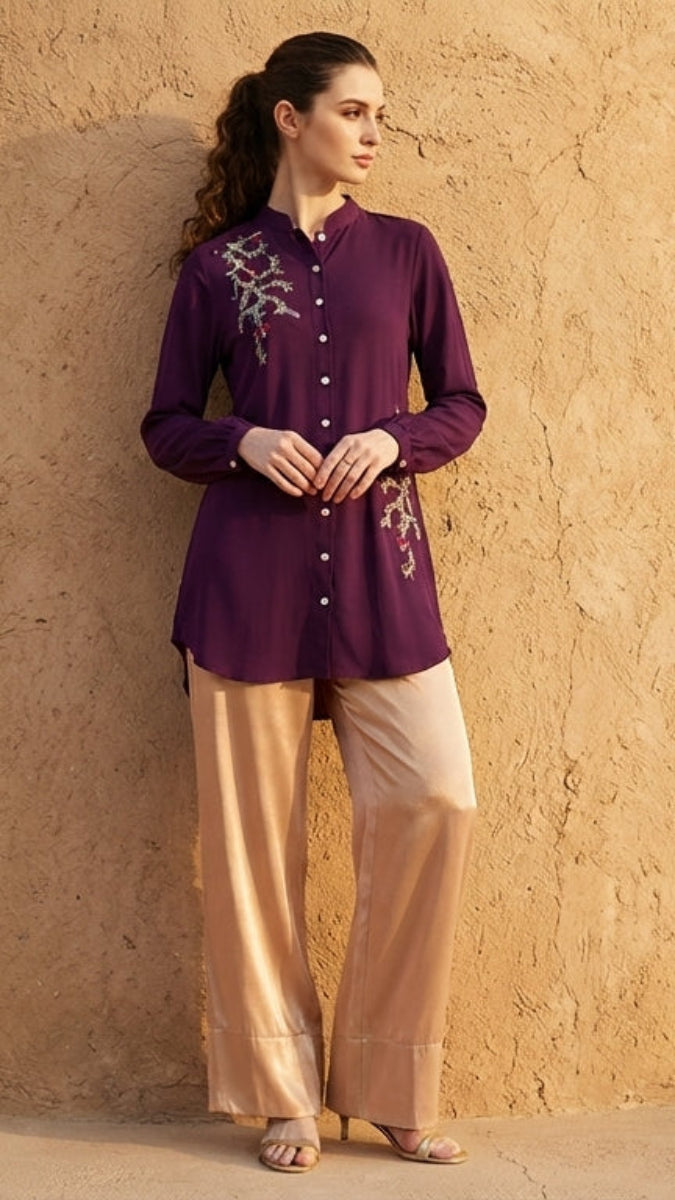 Woman in a purple top and beige pants standing against a beige wall.