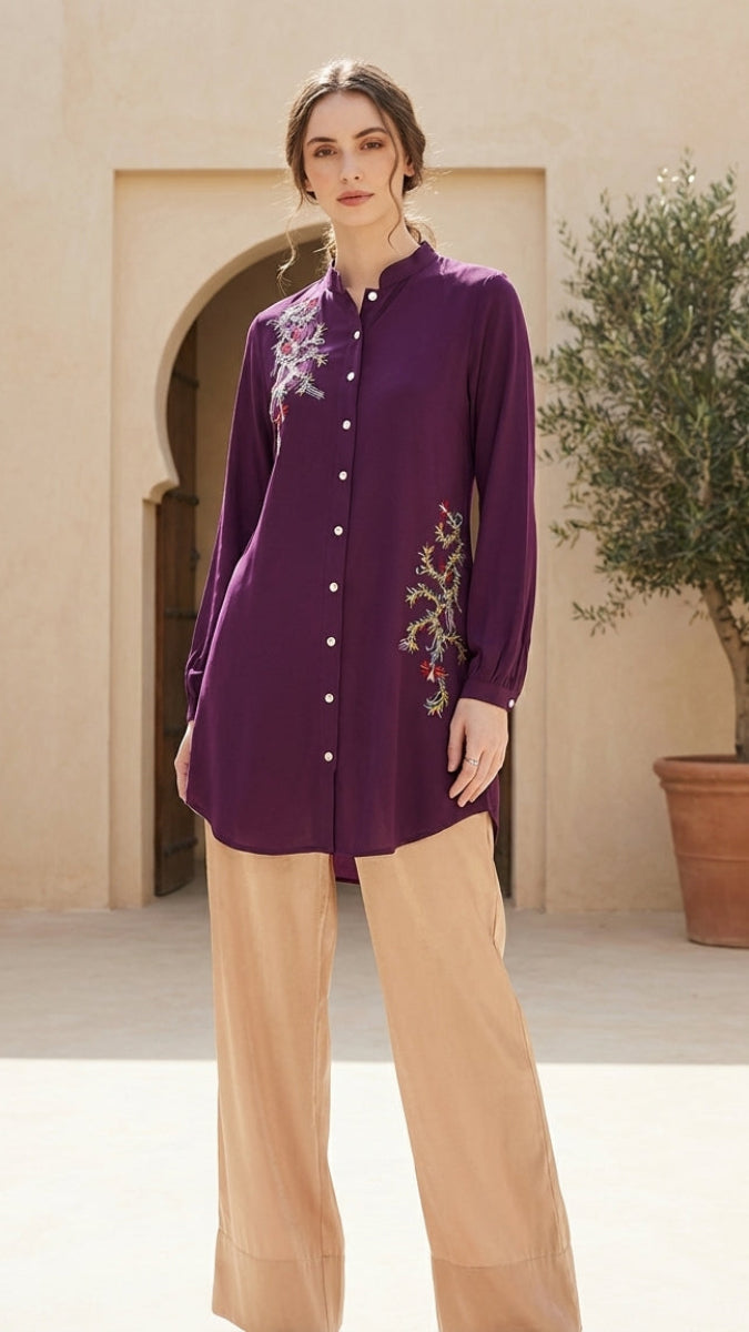 Woman wearing a purple embroidered shirt and beige pants in a decorative setting with a plant.