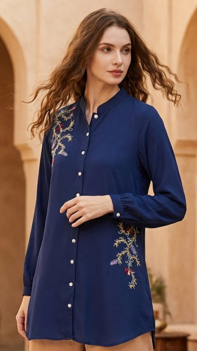 Woman wearing a blue embroidered shirt and beige pants in an outdoor setting with arches and lanterns.