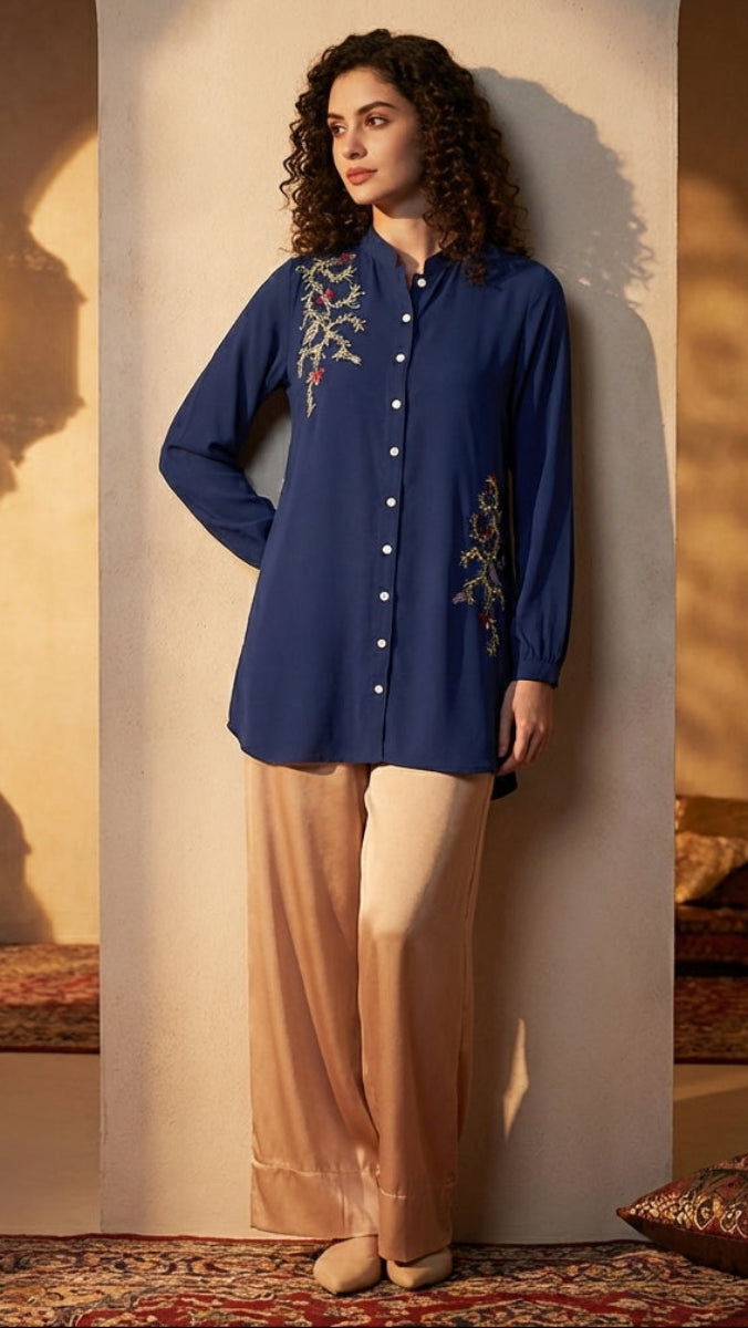 Woman wearing a blue embroidered shirt and beige pants in a warm, decorated room.