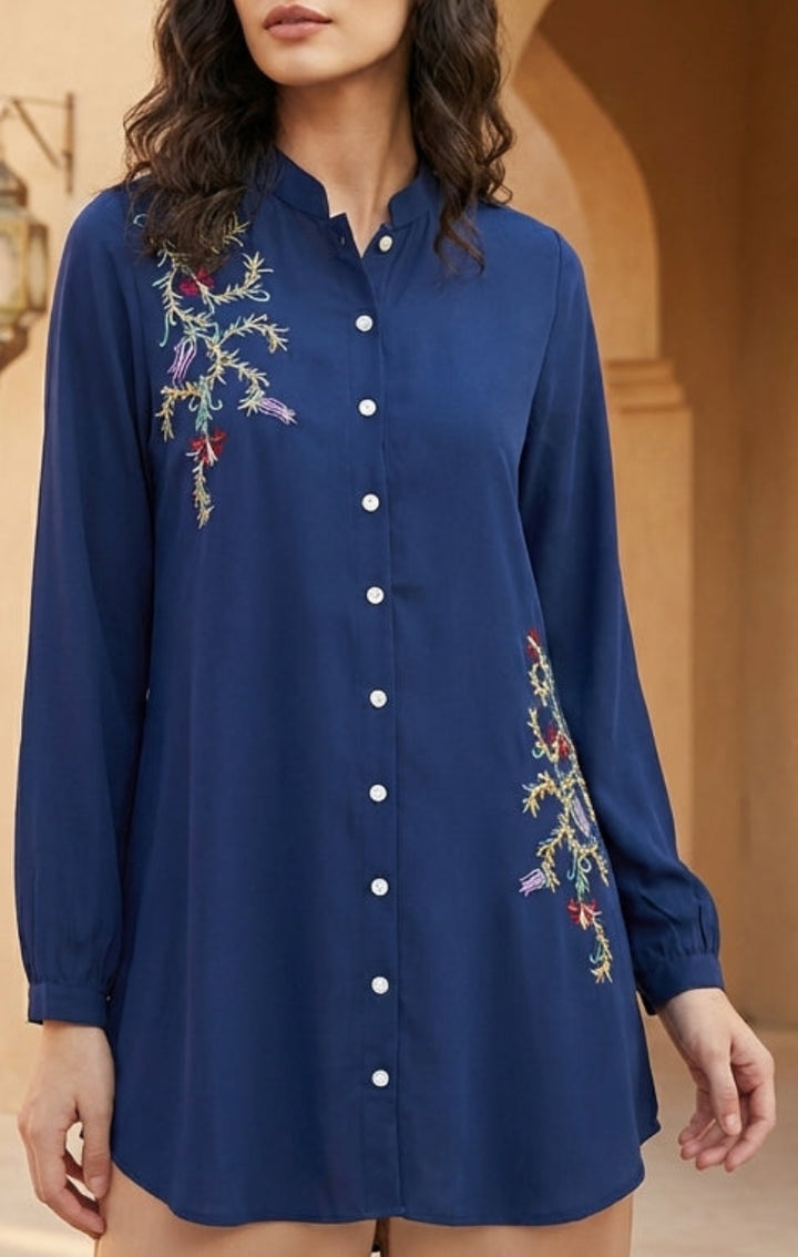 Woman wearing a blue embroidered dress in an outdoor setting with architectural elements.