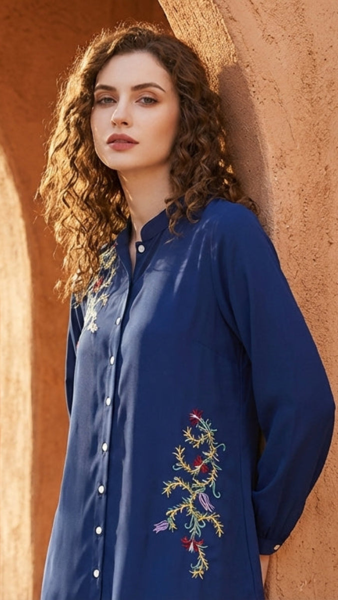 Woman in a blue embroidered shirt standing in front of a stone building with an archway.