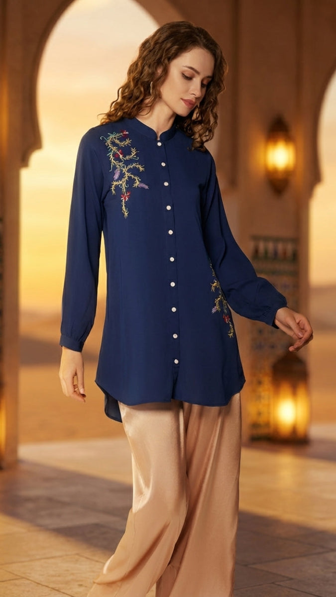 Woman wearing a blue embroidered shirt with beige pants in a warm-toned architectural setting.