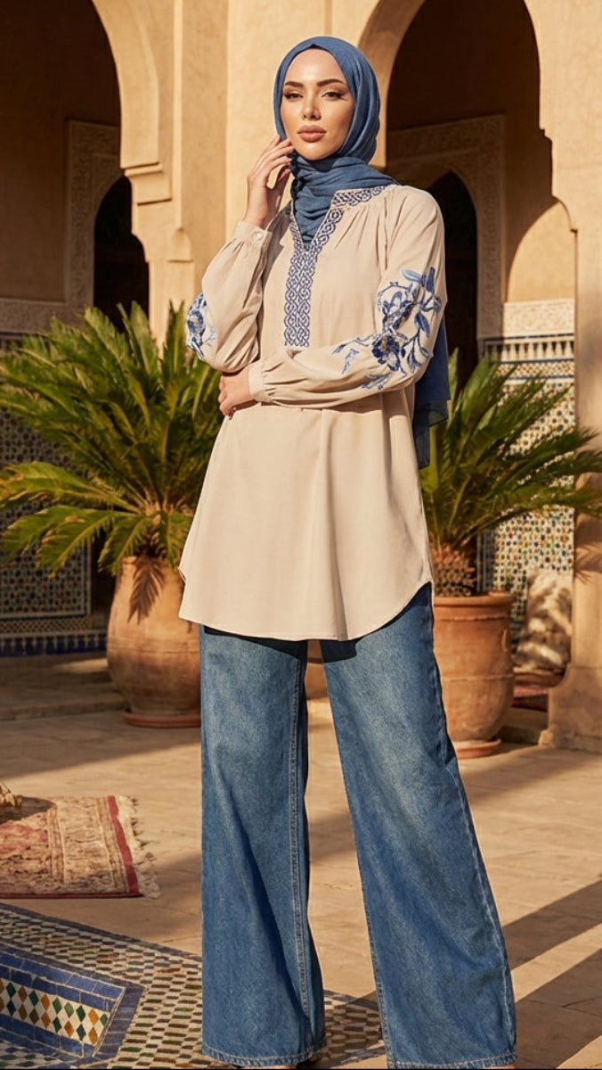 Woman wearing a beige top with blue patterns and jeans, standing in a sunlit outdoor setting with plants and architectural elements.