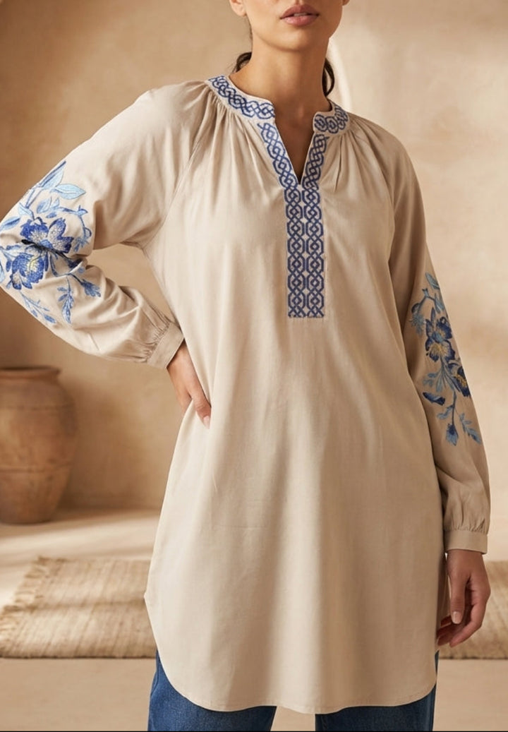 Woman wearing a beige long-sleeve top with blue embroidery in a warm-toned interior setting.