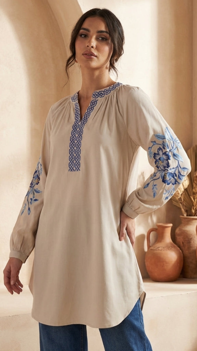 Woman wearing a beige tunic with blue embroidery in a warm-toned indoor setting.