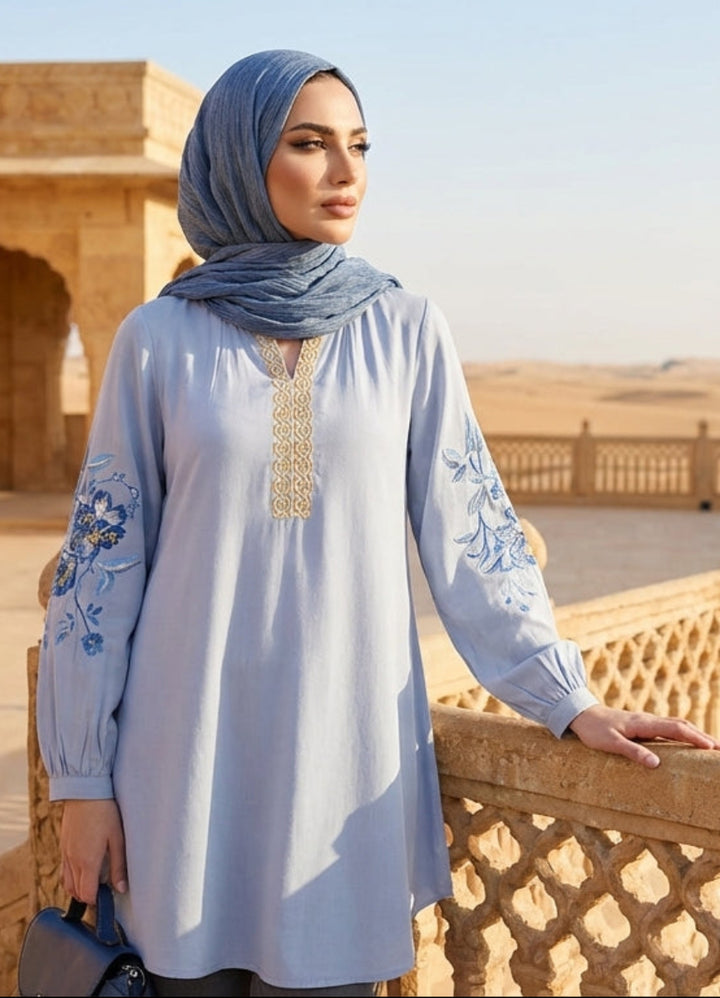 Woman wearing a light blue embroidered dress with a hijab, standing on a balcony with a desert landscape in the background.