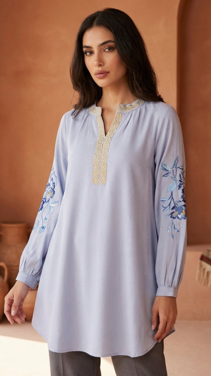Woman wearing a light blue embroidered tunic against a warm-toned wall.