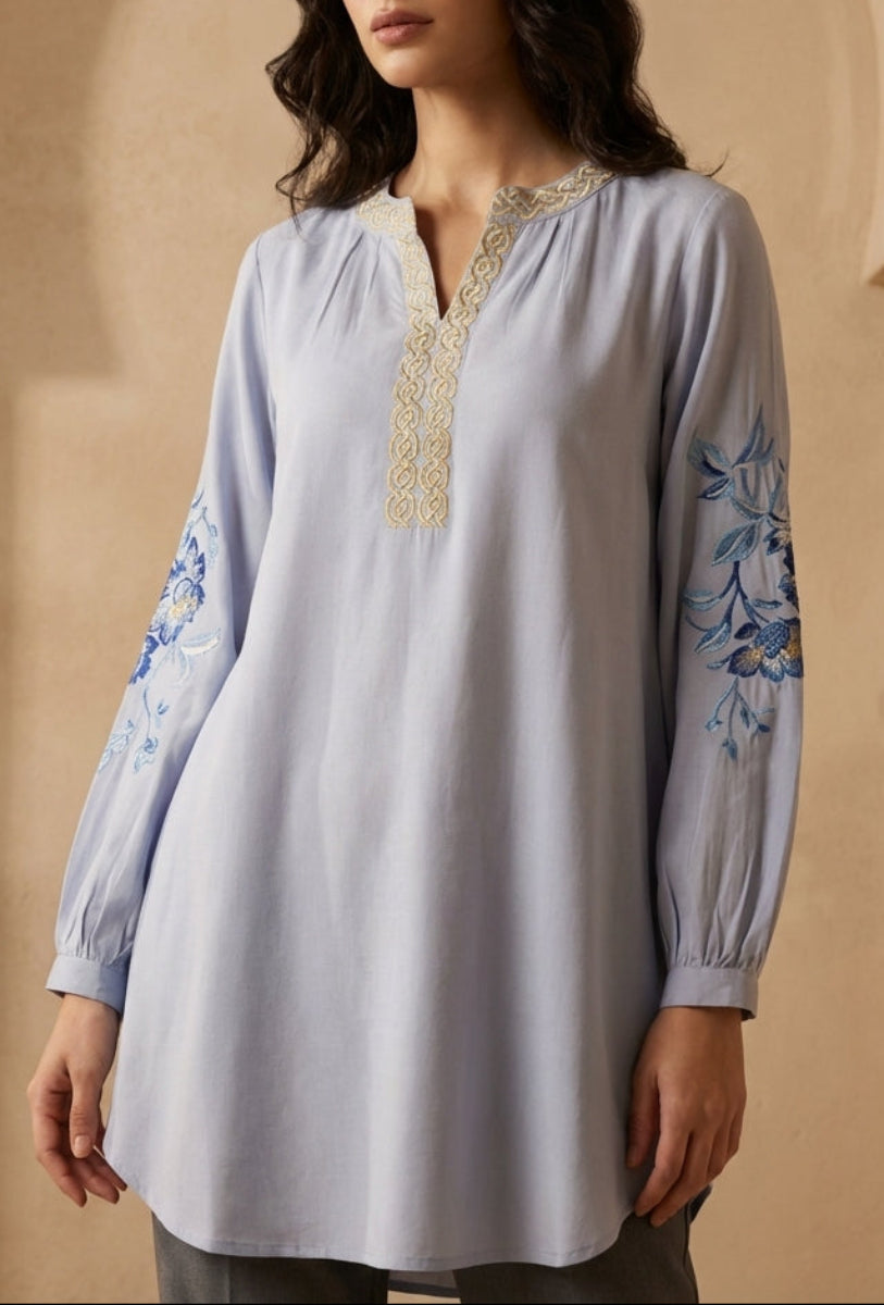 Woman wearing a light blue embroidered top against a beige wall.