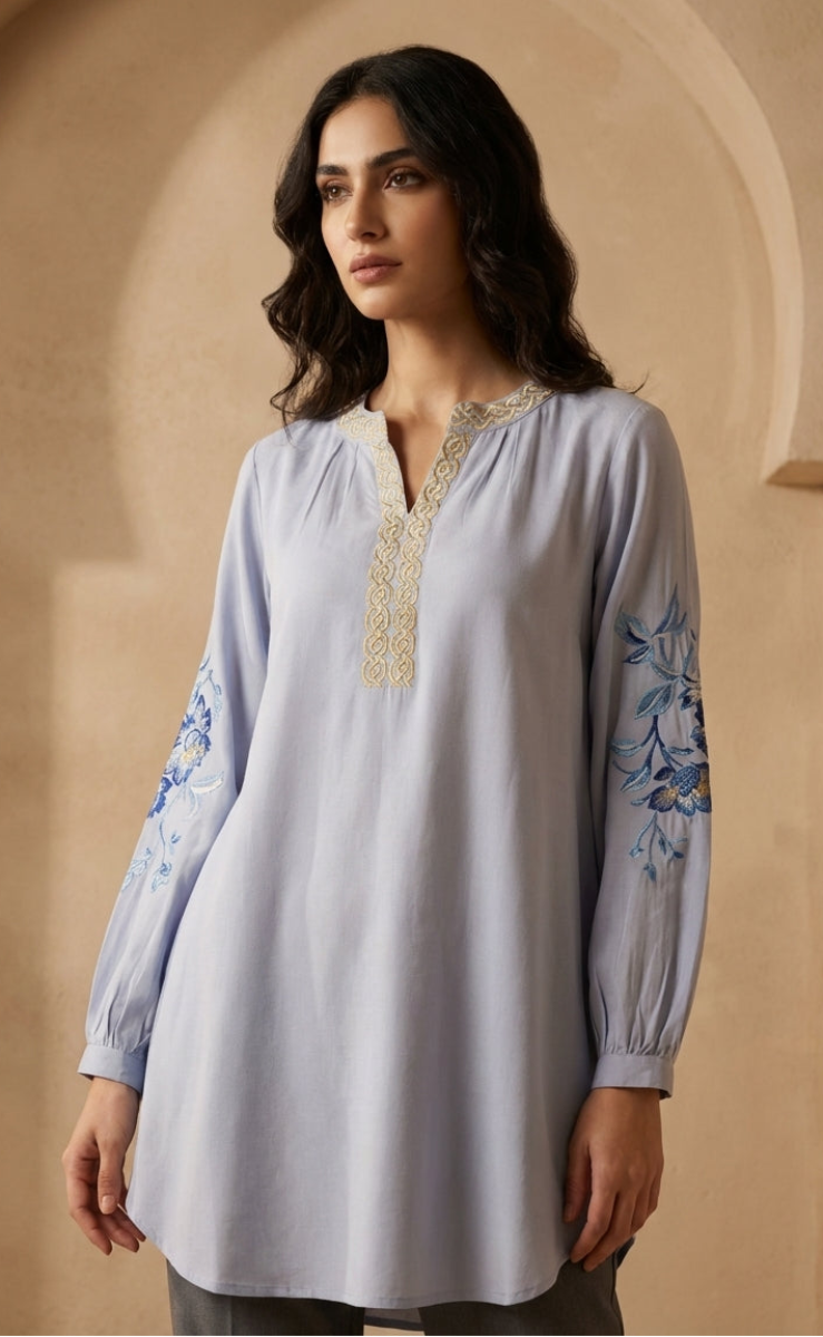 Woman wearing a light blue embroidered top against a beige wall.