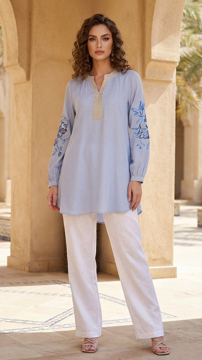 Woman in a light blue embroidered top and white pants standing in a courtyard with palm trees.