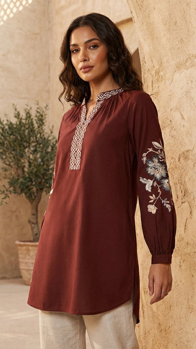 Woman wearing a maroon embroidered top in an outdoor setting with beige walls and plants.