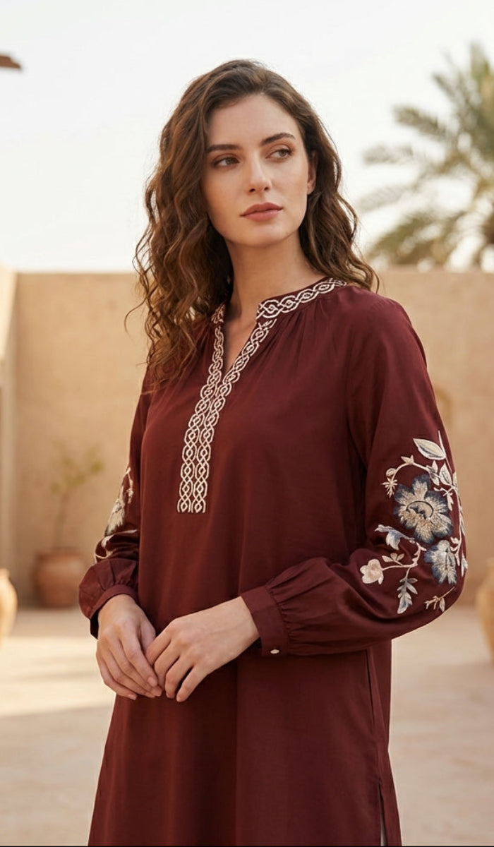 Woman in a burgundy dress with floral embroidery standing in a sunlit courtyard.