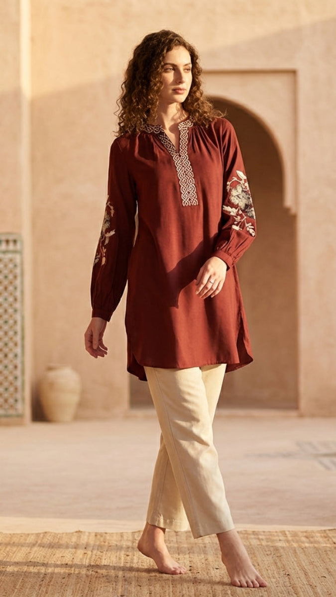 Woman wearing a red embroidered top and white pants standing in a sunlit courtyard with arches.