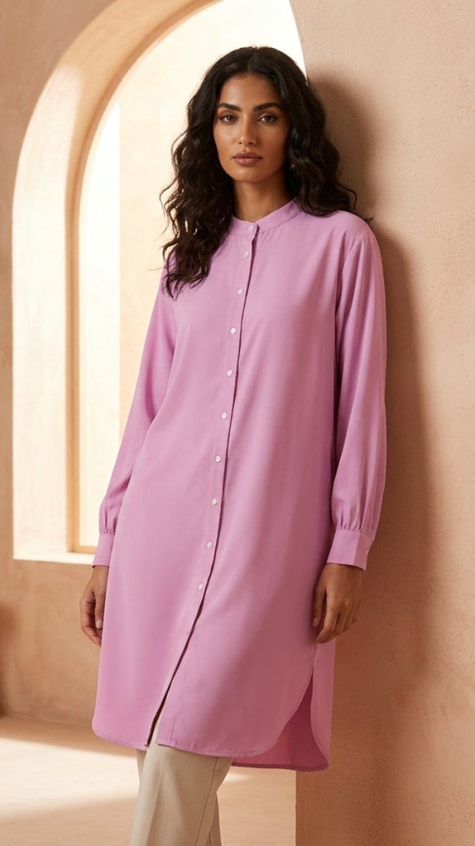 Woman wearing a pink dress standing against a beige wall with a plant and stool in the background.
