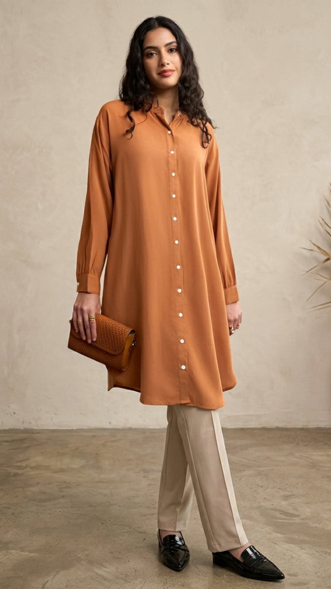Woman wearing a long brown shirt and beige pants standing against a neutral wall with a vase of dried plants.