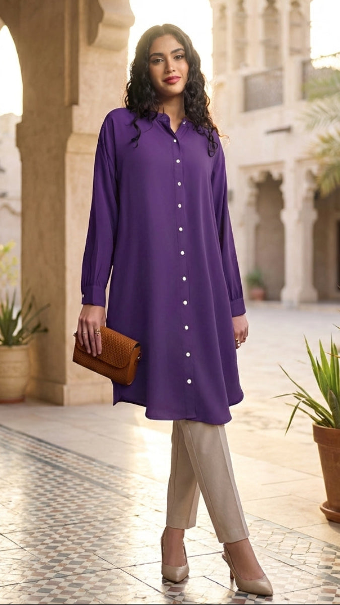 Woman wearing a purple shirt and beige pants standing in an outdoor setting with architectural elements and plants.
