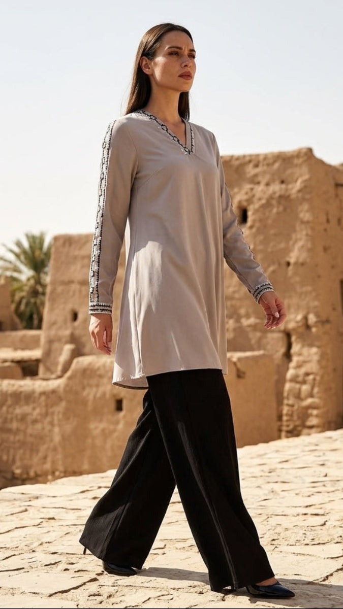 Woman in a long beige dress and black pants walking in front of desert architecture.