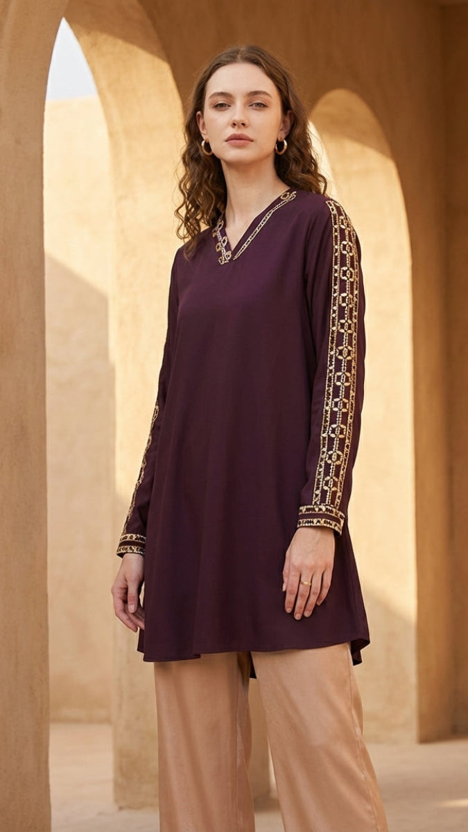 Woman wearing a dark purple top with gold embroidery and beige pants standing in front of stone arches.