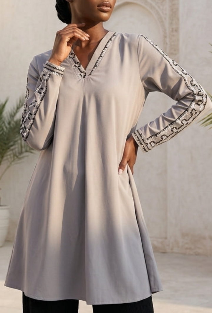 Woman wearing a light gray long-sleeve top with black pants in an outdoor setting with plants and stone architecture.