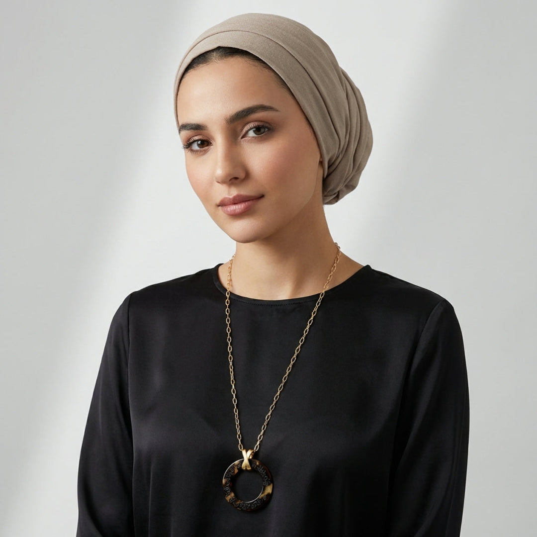 Woman wearing a beige headscarf and black top with a long arabic calligraphy engraved  necklace on a light gray background