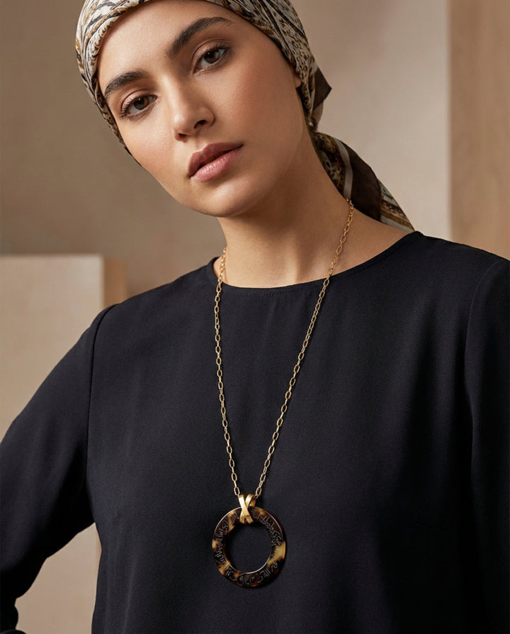Woman wearing a black top and a gold arabic calligraphy engraved  necklace with a large pendant against a neutral background