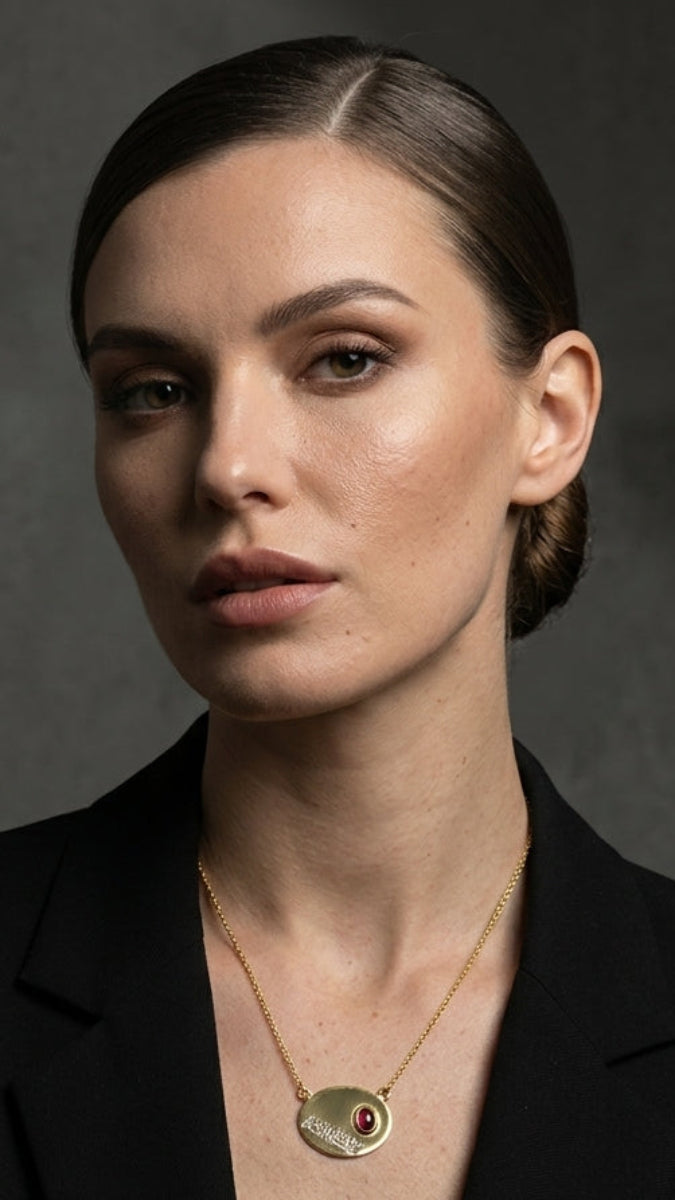 Woman wearing a gold necklace with a red stone against a dark background