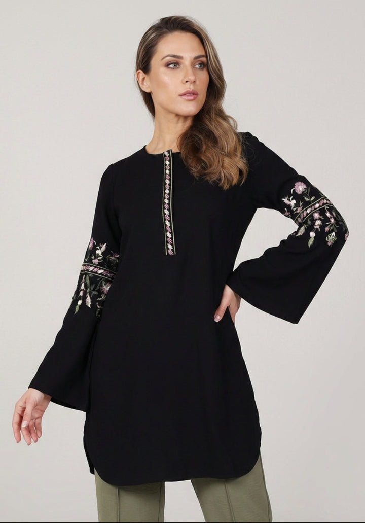 Woman wearing a black long-sleeve top with floral embroidery on a plain background