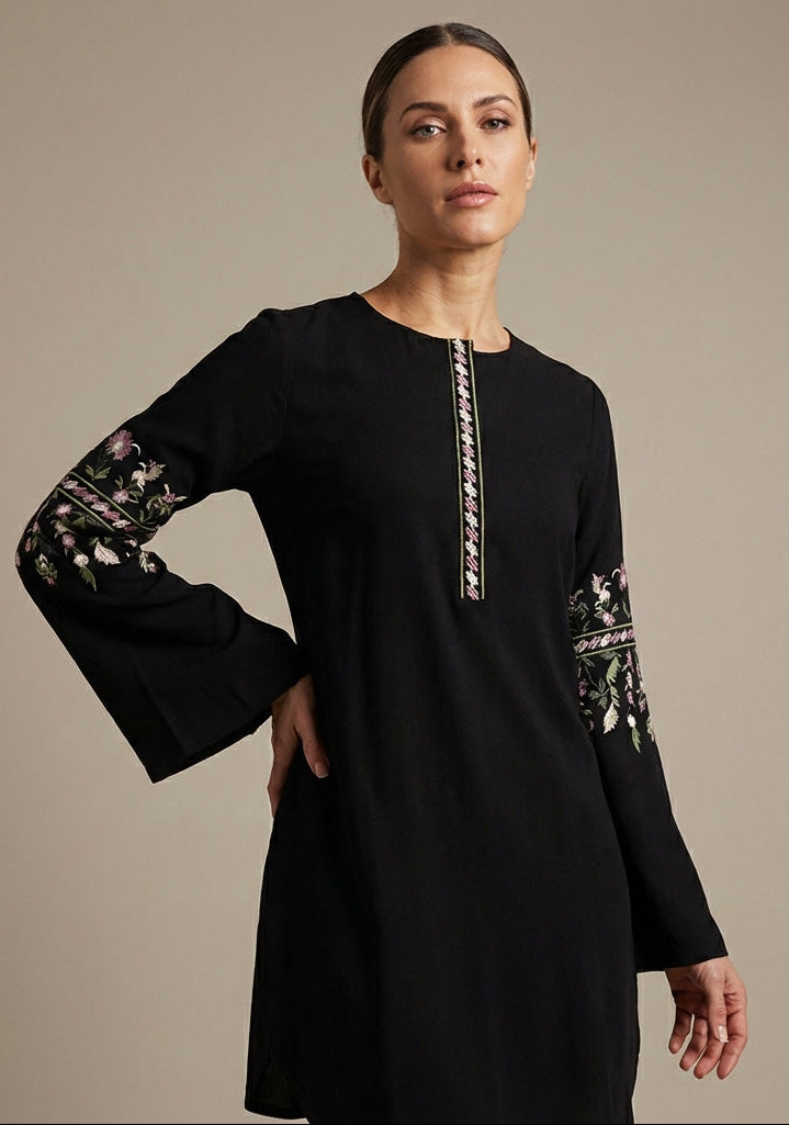 Woman wearing a black dress with floral embroidery on sleeves against a beige background