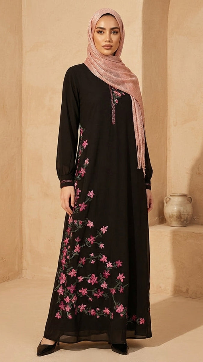 Woman wearing a black dress with pink floral embroidery and a pink hijab in a sandy setting.