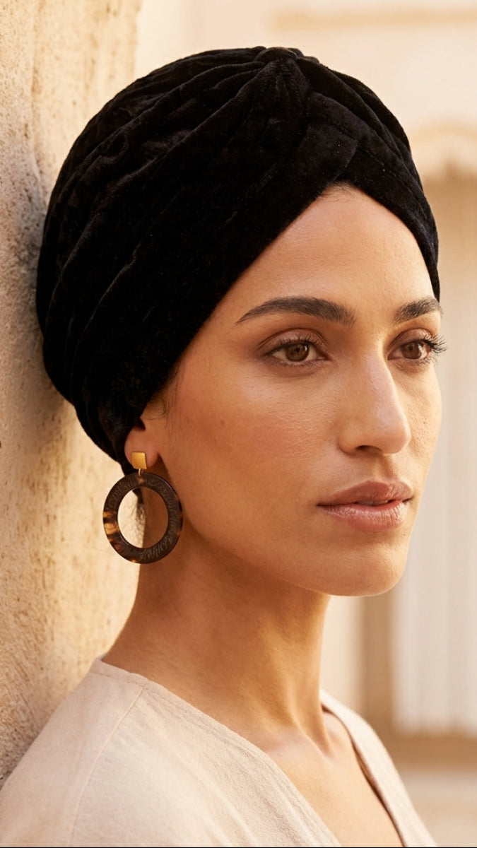 Woman wearing a black turban and large earrings against a beige wall.